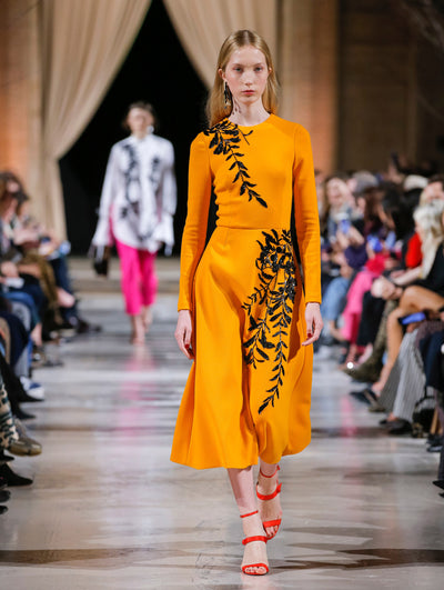The model walks the runway in an orange long-sleeve cocktail dress with black sequin-embroidered leaves. Behind her, the blurred background shows the audience and another model in a white, black and pink ensemble walking the runway.