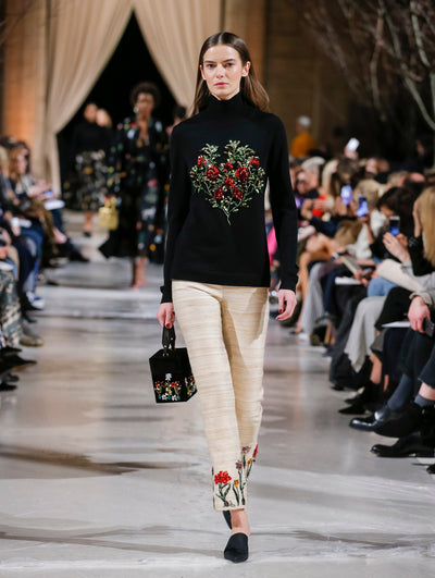 Holding a black cube-like handbag with multi-colored crystal-embroidered florals, the model walks the runway in a black long-sleeve turtleneck pullover with multi-colored sequin-embroidered florals and beige trousers with multi-colored sequin-embroidered florals. Behind her, the blurred background shows the audience and several models walking the runway.