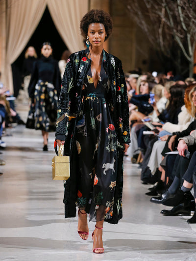 Holding a gold cube-like purse, the model walks the runway in a black open-front long-sleeve velvet coat with a threadwork embroidered animal and floral design, layered over a black V-neck silk dress printed with an all-over animal and floral design. Behind her, the blurred background shows the audience and several models walking the runway in various directions.
