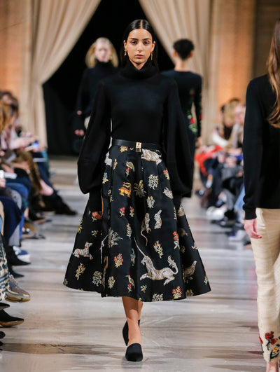 The model walks the runway in a black long balloon sleeve turtleneck pullover and black skirt printed with an all-over animal and floral design, paired with a black and gold belt. Behind her, the blurred background shows the audience and several models walking the runway in various directions.
