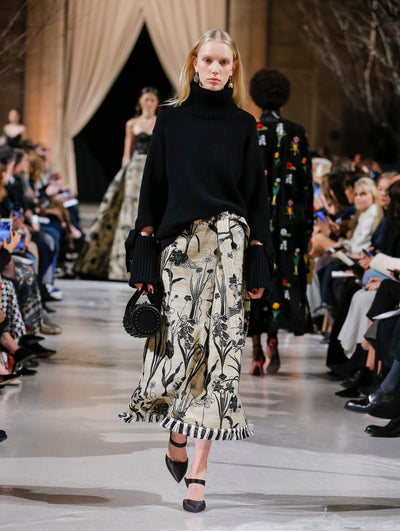 Holding a black tube-like handbag, the model walks the runway in a black long-sleeve turtleneck bow-detailed pullover and beige fringed skirt printed with black florals. Behind her, the blurred background shows the audience and several models walking the runway in various directions.