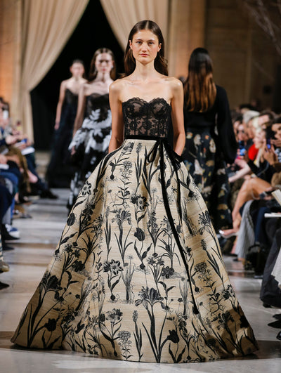The model walks the runway in a strapless ballgown with a black lace bow-detailed bustier bodice and beige voluminous wool skirt printed with black florals. Behind her, the blurred background shows the audience and several models walking the runway in various directions.