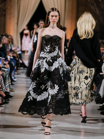 The model walks the runway in a black lace cocktail dress with a bustier bodice and tiered skirt with all-over white threadwork embroidered leaves. Behind her, the blurred background shows the audience and several models walking the runway in various directions.