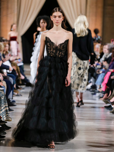 The model walks the runway in a black evening gown with a lace bustier bodice and a tulle scalloped skirt. Behind her, the blurred background shows the audience and several models walking the runway in various directions.
