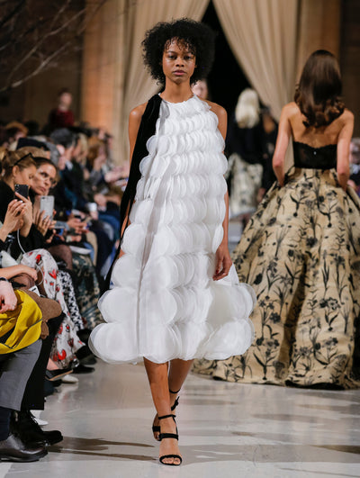 The model walks the runway in a white sleeveless bow-detailed cocktail dress made entirely of scalloped pleating. Behind her, the blurred background shows the audience and several models walking the runway in various directions.