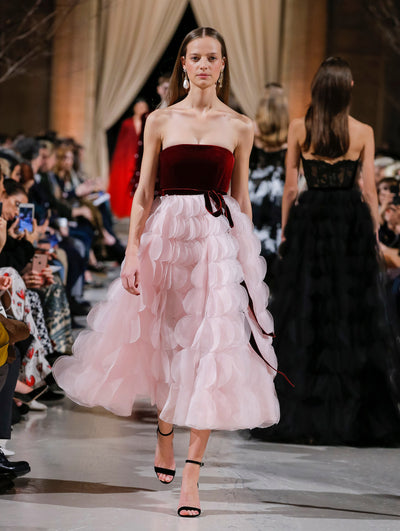 The model walks the runway in a strapless cocktail dress with a burgundy velvet bow-detailed bodice and a skirt made entirely of scalloped pleating. Behind her, the blurred background shows the audience and several models walking the runway in various directions.