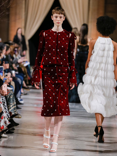 The model walks the runway in a burgundy bow-detailed long-sleeve velvet jacket with crystal embroidery and a coordinating burgundy velvet skirt with crystal embroidery. Behind her, the blurred background shows the audience and several models walking the runway in various directions.
