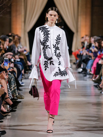 Holding a maroon gardenia appliqué handbag, the model walks the runway in a white button-up long-sleeve blouse with black sequin-embroidered sleeves. Behind her, the blurred background shows the audience.