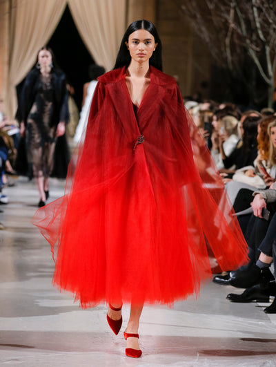The model walks the runway in an ombré long-sleeve tulle opera coat where shades of deep burgundy transition to bright red with a crystal flower pin closure. Behind her, the blurred background shows the audience and several models walking the runway in various directions.