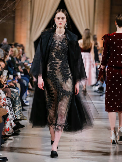 The model walks the runway in a black long-sleeve tulle opera coat, layered over a black semi-sheer mesh dress with threadwork embroidered leaves and fringe trim. Behind her, the blurred background shows the audience and several models walking the runway in various directions.