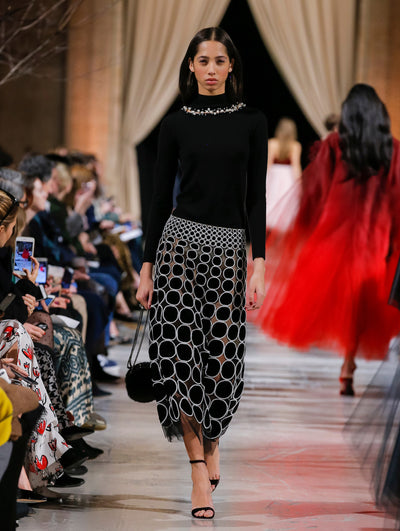 Holding a black billiards bag, the model walks the runway in a black long-sleeve mock-neck pullover with crystal embroidery concentrated at the neckline and a black skirt with all-over circle crystal embroidery on a semi-sheer tulle base. Behind her, the blurred background shows the audience and several models walking the runway in various directions.