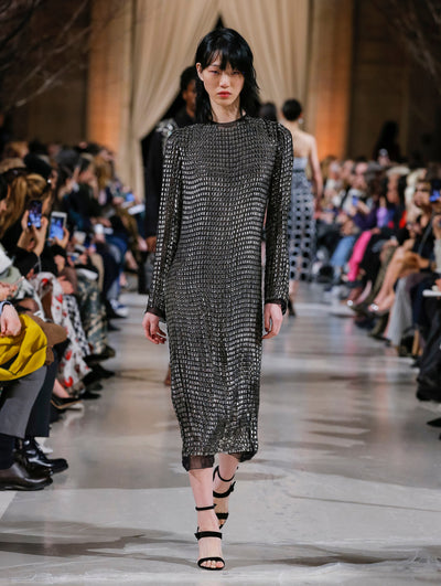 The model walks the runway in a black long-sleeve cocktail dress with all-over metallic grid detailing. Behind her, the blurred background shows the audience and several models walking the runway in various directions.
