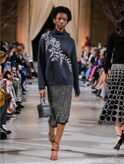 Holding a silver cube-like handbag, the model walks the runway in a gray long-sleeve turtleneck pullover with crystal-embroidered leaves and a black skirt with all-over metallic grid detailing. Behind her, the blurred background shows the audience and several models walking the runway in various directions.