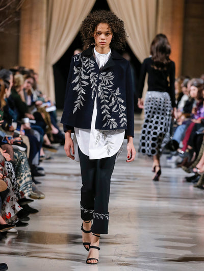 The model walks the runway in a navy long-sleeve wool coat with crystal-embroidered leaves, layered over a white button-up blouse with silver sequin-embroidered leaves and black trousers with crystal-embroidered cuffs. Behind her, the blurred background shows the audience and several models walking the runway in various directions.