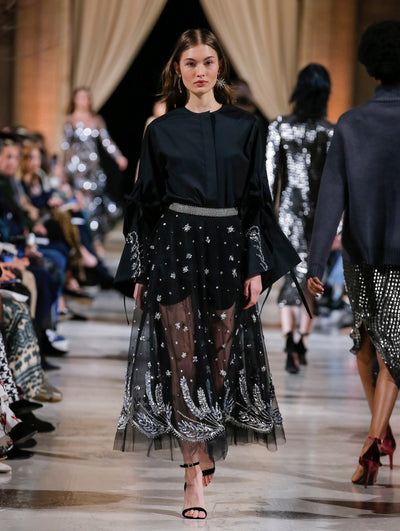 The model walks the runway in a navy long-sleeve button-up blouse with silver sequin-embroidered cuffs, layered over a black semi-sheer tulle skirt with all-over sequin embroidery. Behind her, the blurred background shows the audience and several models walking the runway in various directions.