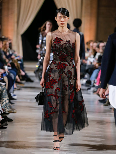Holding a black crystal-embroidered gardenia appliqué handbag, the model walks the runway in a black sleeveless illusion-neck cocktail dress with red sequin-embroidered florals. Behind her, the blurred background shows the audience and several models walking the runway in various directions.
