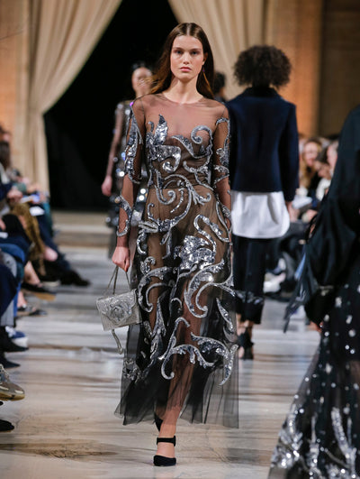 Holding a silver crystal-embroidered gardenia appliqué handbag, the model walks the runway in a black long-sleeve illusion-neck cocktail dress with silver sequin-embroidered florals. Behind her, the blurred background shows the audience and several models walking the runway in various directions.