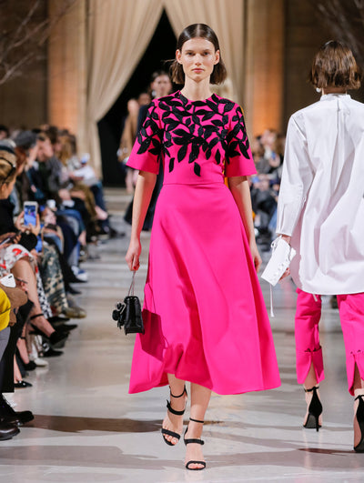 Holding a black gardenia appliqué handbag, a model walks the runway in a pink short-sleeve cocktail dress with black sequin-embroidered leaves concentrated on the bodice. Behind them, the blurred background shows the audience and several models walking the runway in various directions.