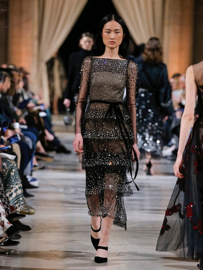 The model walks the runway in a black long-sleeve boat-neck cocktail dress with all-over crystal embroidery and a tiered bow-detail skirt. Behind her, the blurred background shows the audience and several models walking the runway in various directions.