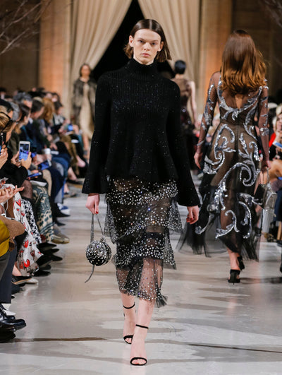 Holding a black crystal-embroidered billiards bag, the model walks the runway in a black long-sleeve ribbed knit turtleneck pullover and black semi-sheer tulle tiered skirt with all-over crystal embroidery. Behind her, the blurred background shows the audience and several models walking the runway in various direction
