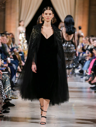 The model walks the runway in a black long-sleeve tulle opera coat with gold sequin embroidery, layered over a black V-neck cocktail dress. Behind her, the blurred background shows the audience and several models walking the runway in various directions.