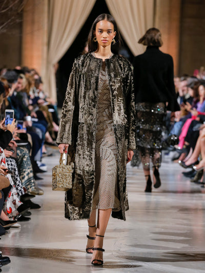 Holding a gold sequin-embroidered cube-like handbag, the model walks the runway in a metallic brown open-front long-sleeve fur coat, layered over a metallic semi-sheer mesh dress with threadwork embroidered leaves. Behind her, the blurred background shows the audience and several models walking the runway in various directions.