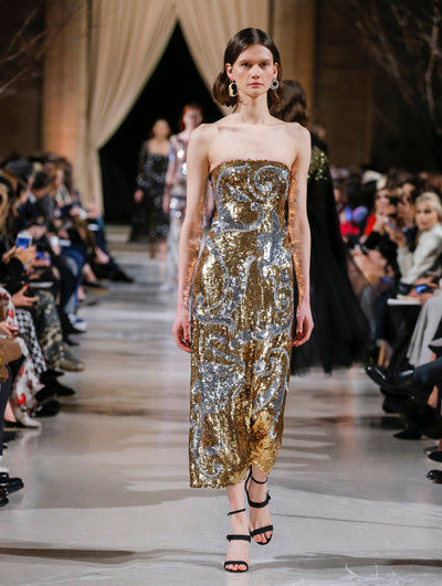 The model walks the runway in a strapless cocktail dress with all-over silver and gold swirling sequin embroidery. Behind her, the blurred background shows the audience and several models walking the runway in various directions.