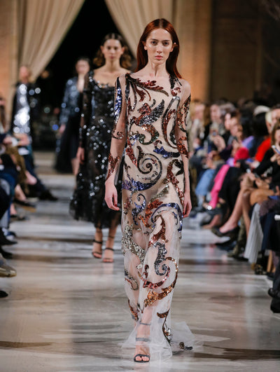 The model walks the runway in a white semi-sheer illusion-neck long-sleeve evening gown with swirling multi-colored sequin embroidery. Behind her, the blurred background shows the audience and several models walking the runway.
