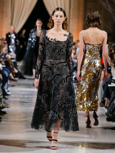 The model walks the runway in a black semi-sheer illusion-neck long-sleeve cocktail dress with swirling black and silver embroidery. Behind her, the blurred background shows the audience and several models walking the runway in various directions.