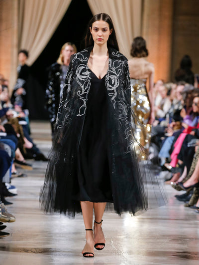 The model walks the runway in a black open-front long-sleeve tulle opera coat with a sequin-embroidered design, layered over a black V-neck cocktail dress. Behind her, the blurred background shows the audience and several models walking the runway in various directions.