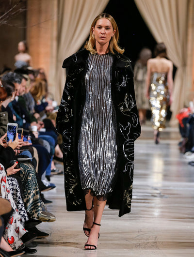 The model walks the runway in a black open-front long-sleeve fur coat with a sequin-embroidered design, layered over a black all-over silver sequin-embroidered cocktail dress. Behind her, the blurred background shows the audience and several models walking the runway in various directions.