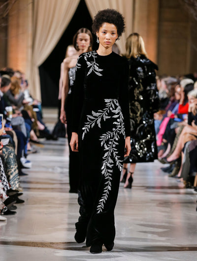 The model walks the runway in a black long-sleeve column evening gown with crystal-embroidered leaves. Behind her, the blurred background shows the audience and several models walking the runway in various directions.