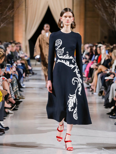 The model walks the runway in navy long sleeve cocktail dress with white curling vine embroidery. Behind her, the blurred background shows the audience and another model in a brown ensemble walking the runway.