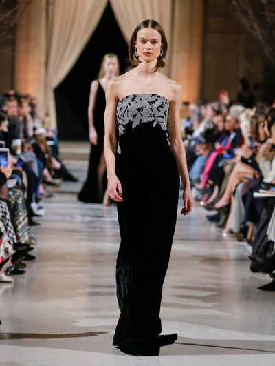 The model walks the runway in a black column evening gown with crystal-embroidered leaves concentrated on the bodice. Behind her, the blurred background shows the audience and a model walking the runway in a black emsemble.
