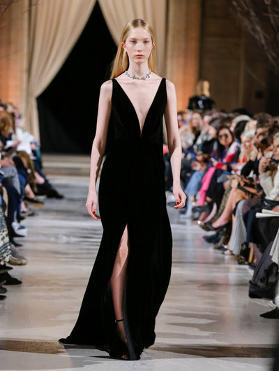 The model walks the runway in a black V-neck evening gown with a high front slit. Behind her, the blurred background shows the audience.