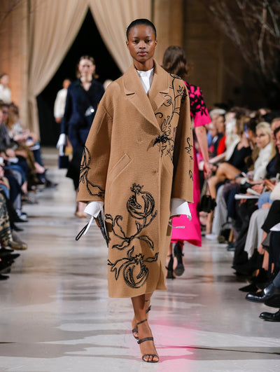 Holding a thin black clutch, the model walks the runway in a brown long-sleeve oversized wool coat with a crystal leaf pin closure and black pomegranate embroidery, layered over a white long-sleeve blouse. Behind her, the blurred background shows the audience and several models walking the runway in various directions.