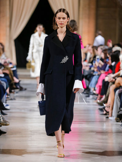 Holding a navy cube-like handbag, the model walks the runway in a navy long-sleeve oversized wool coat with a crystal leaf pin closure, layered over a white long-sleeve blouse. Behind her, the blurred background shows the audience and several models walking the runway in various directions.