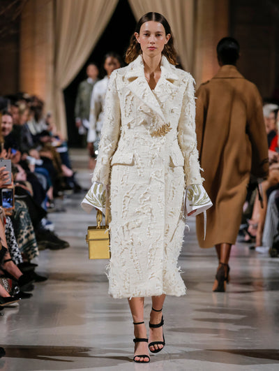 Holding a gold cube-like handbag, the model walks the runway in an ivory textured wool coat with a crystal leaf pin closure, layered over a white long-sleeve blouse with gold threadwork embroidered cuffs. Behind her, the blurred background shows the audience and several models walking the runway in various directions.