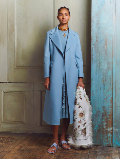 A model wears a light blue coat and is holding a fur scarf with print