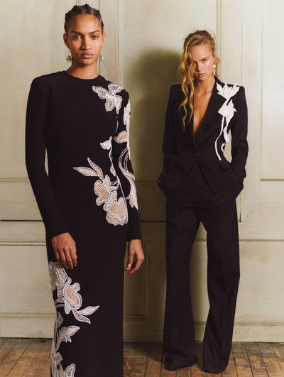 Two models; the model wears a black dress with white flowers embroidered and the model on the right wears a black suit set with white flowers embroidered