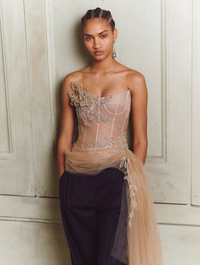 A model wears a nude tulle top with flowers and ivy embroidered with black pants