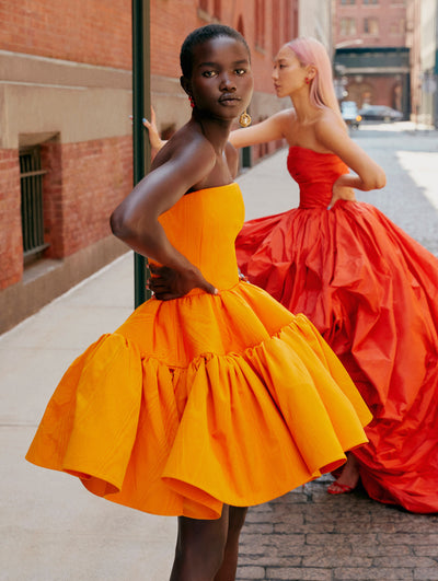 Two models are seen posing on the New York city sidewalk, one wearing a bright orange Oscar de la Renta cocktail dress and the other wearing wearing a red Oscar de la Renta gown. 