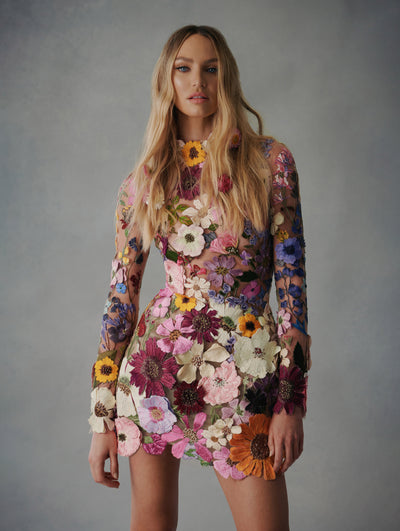 The model wears a long sleeve mini dress with flowers embroidered all over, the flowers range and come in yellow, white, and pink.