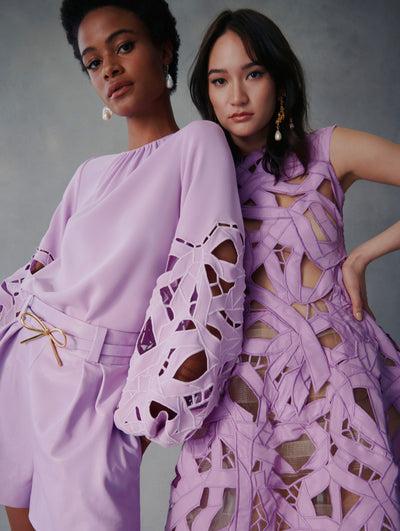Two models pose in lavender Fall 2021 Looks. The model in the left is wearing lavender shorts with a belt that has a bow in the center, paired with a lavender blouse with bow cutouts in the sleeve. The model to the right is wearing a dress with the same bow cutouts all around.