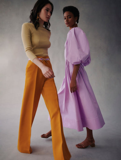 Two models pose in Fall 2021 Looks. The model in the front is wearing orange pants with a lilac bow belt and beige sweater. The model in the back is wearing a lilac poplin cotton dress with puff sleeves.