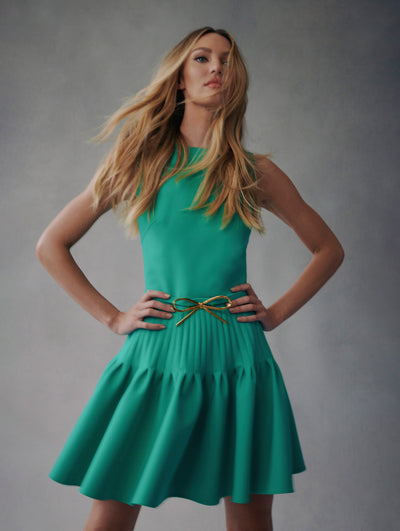 The model wears an aquamarine, sleeveless, mini dress with a bow detail in the belt and ruffle skirt from Fall 2021 
