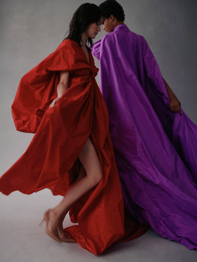 Two models wear gowns from Fall 2021. They are turning and one wears a purple gown with a cape and the model in the left wears a red gown.