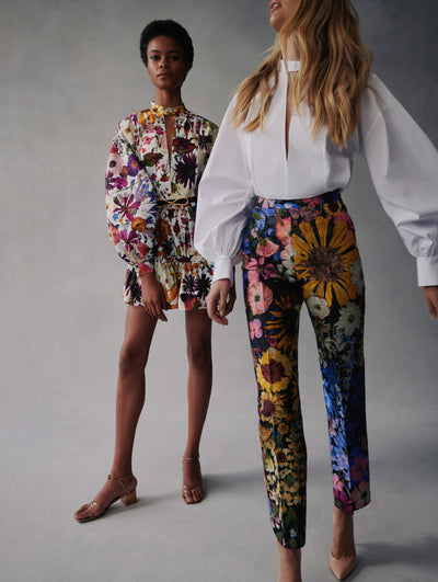 Two models pose in Fall 2021 Looks. The model in the right wear's floral pants with a white blouse with puff sleeves. The model standing behind her to the left wear's floral shorts with a matching blouse.