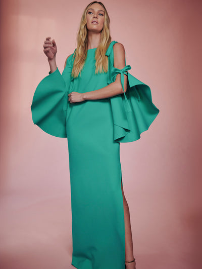 The model poses with a pink backdrop wearing an aquamarine long dress with ruffle open sleeves with bow details from Fall 2021