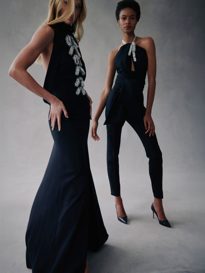 Two models pose in Fall 2021 Looks. The model in the right is wearing black pants with a black halter blouse with crystal details in the neck. The model in the left is wearing a black halter gown with crystal details in the top.